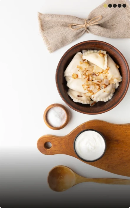 A top-down view of a rustic clay bowl filled with homemade vareniki or pierogi dumplings topped with caramelized onions. Accompaniments include a small bowl of sour cream on a wooden cutting board and a small bowl of salt, all on a clean white background